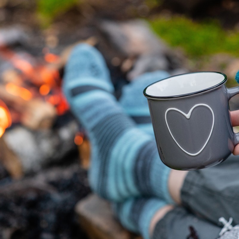 Woman warms feet in socks around bonfire and holds cup of a hot tea