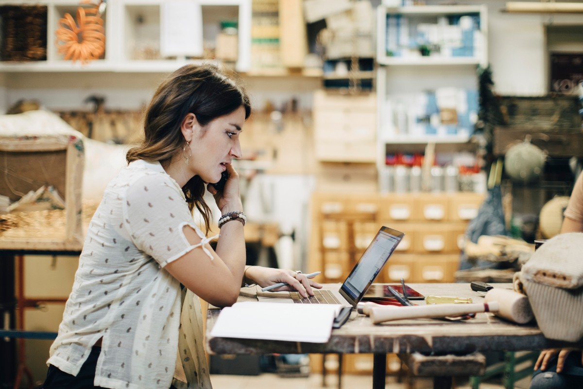 Side view of female upholstery worker using laptop while talking on phone at workbench in workshop