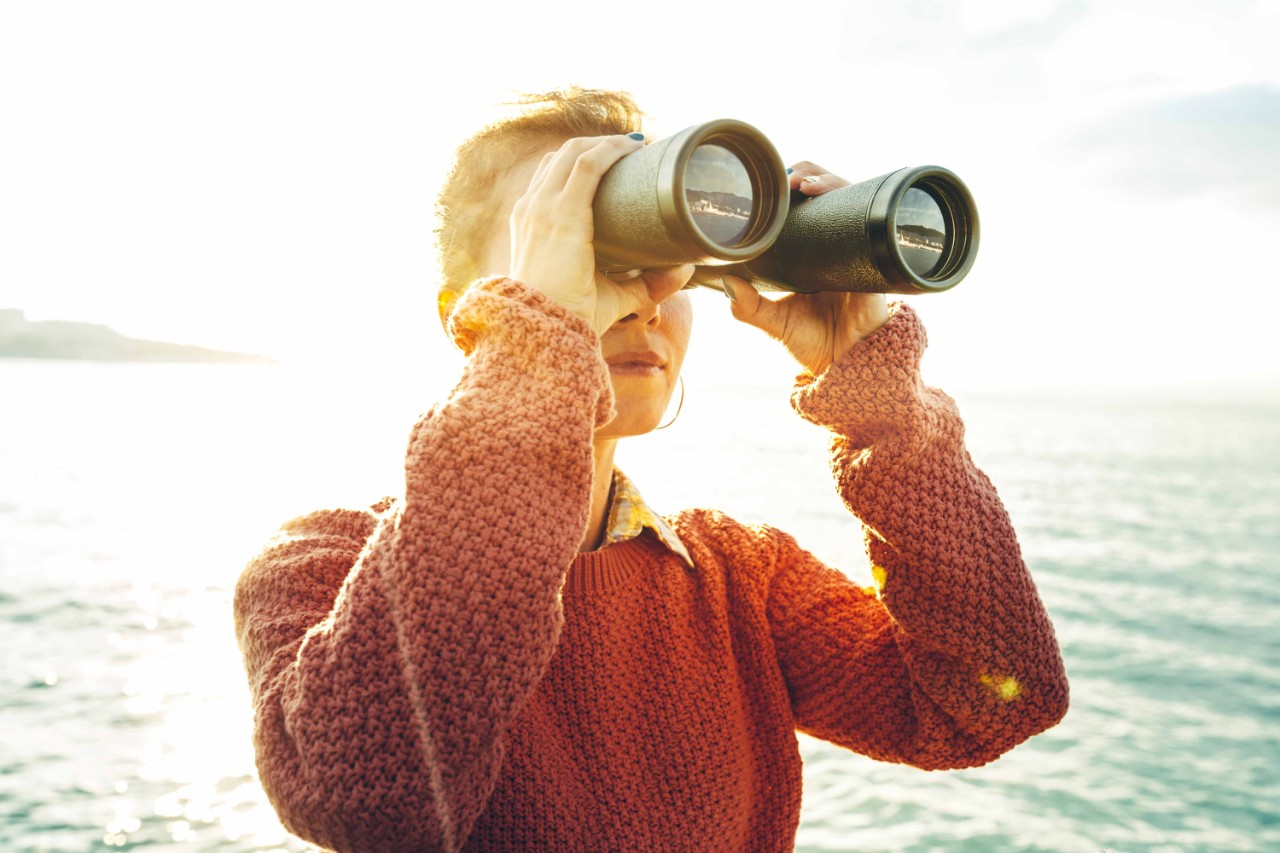 Beautiful young girl in an orange jumper looks through binoculars at the sea on a bright sunny day, low angle