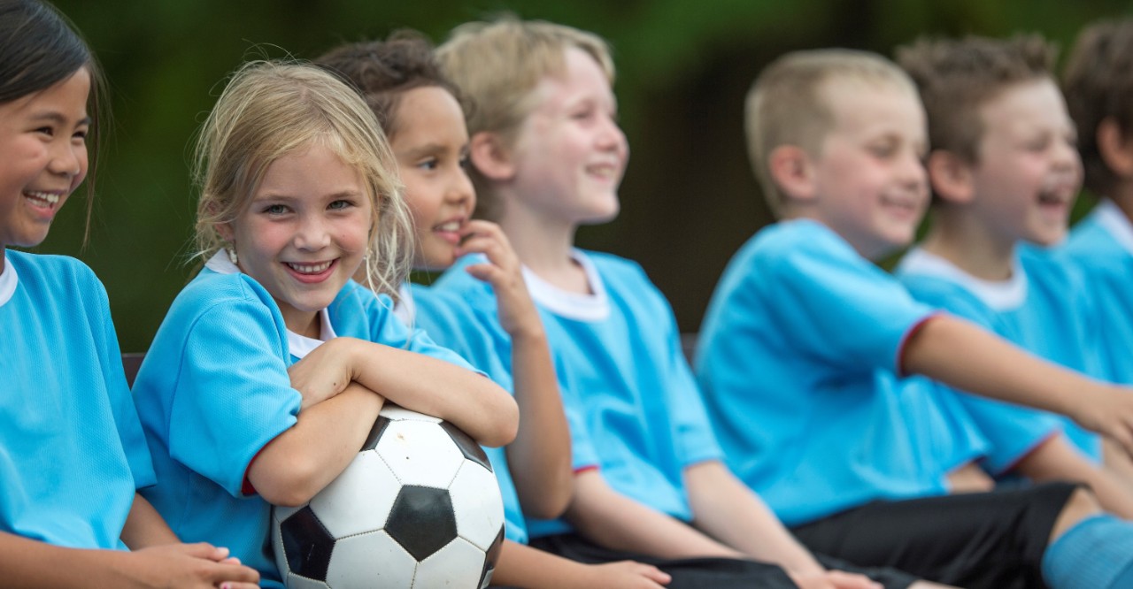 A multi-ethnic group of elementary age children are sitting in a row on the bench and are waiting for the soccer game to start. One girl is holding a soccer ball and is smiling while looking at the camera.