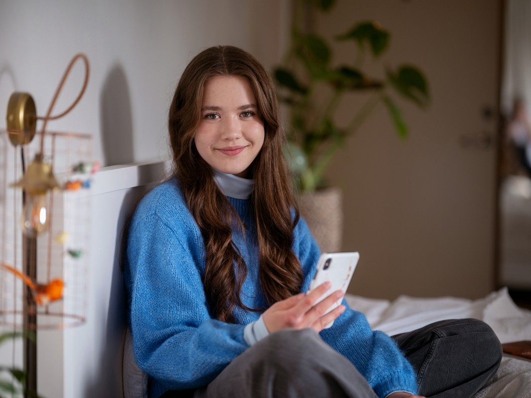 Jente i blå genser sitter i senga og holder en mobil
