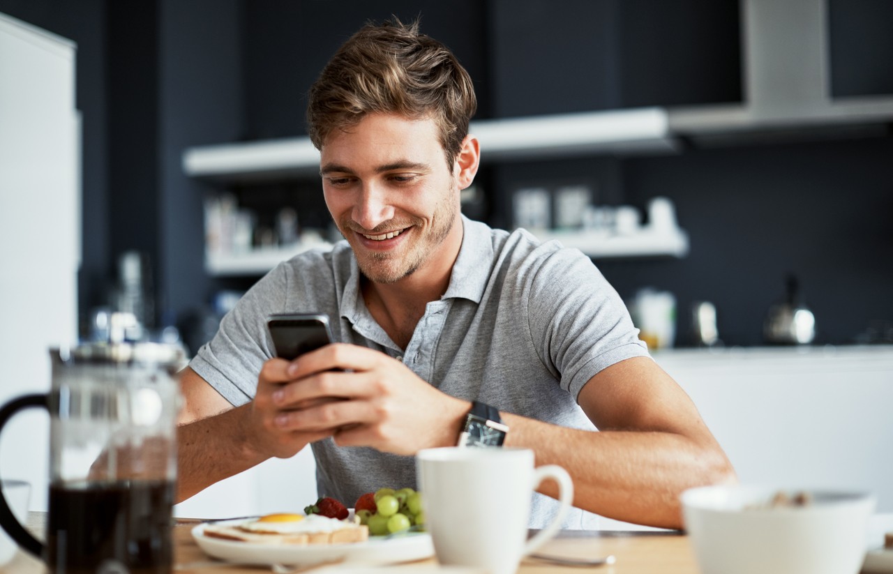 Shot of a happy young man using his cellphone while enjoying breakfast at home