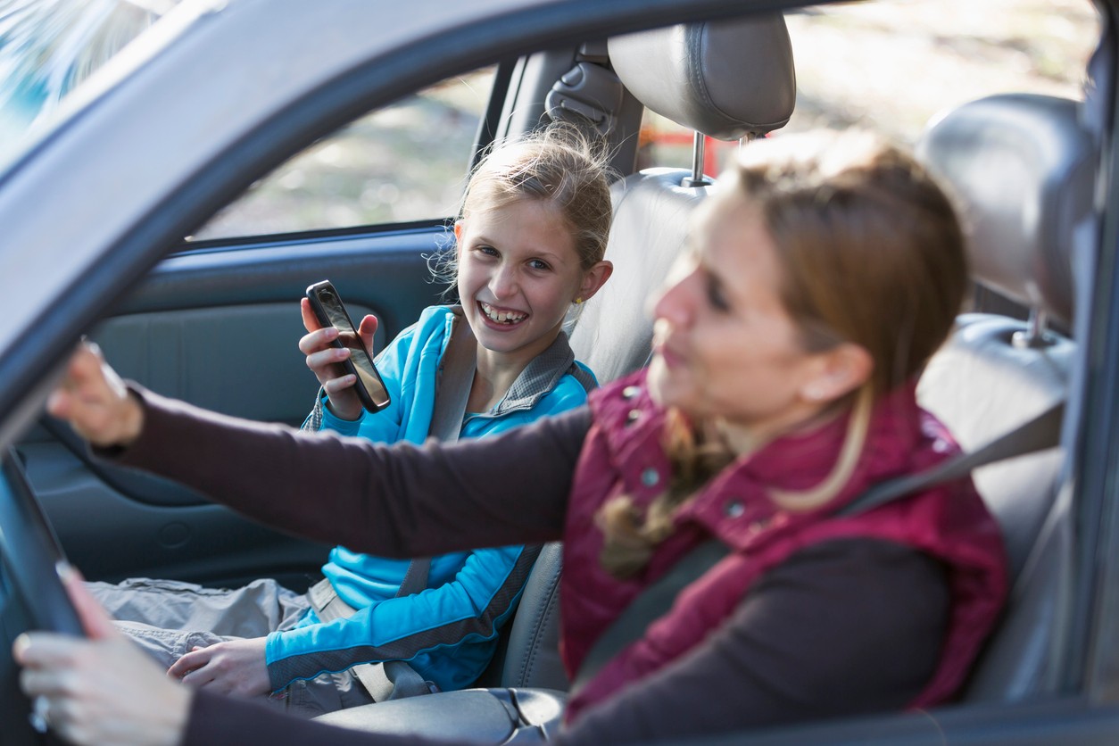 A happy mother and daughter on a road trip, travelling in a car.  The focus is on the little girl, 9 years hold, sitting in the front passenger seat, smiling at her mom, showing her something on her mobile phone.
